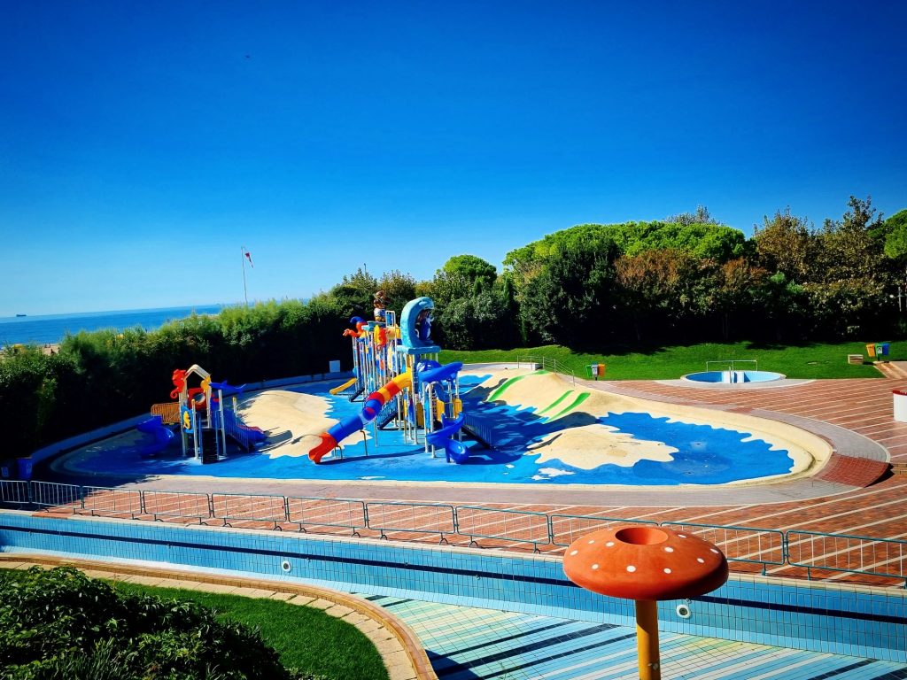 Der Aquapark Mare im Union Lido in Cavallino, Italien, am Ende der Sommersaison. Alle Becken, einschließlich des Strömungskanals, sind vollständig entleert. Zu sehen sind das leere Kinderbecken mit bunten Wasserrutschen und Spielgeräten sowie die abgesperrten Poolbereiche. Im Hintergrund sind Pinien und die Adriaküste zu erkennen.
