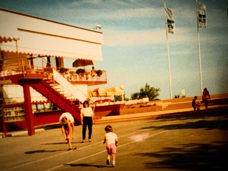 Farbfotografie der Piazza Lido im Union Lido in Cavallino, Italien, aus dem Jahr 1987. Zu sehen sind das damalige Gebäude mit markanter roter Außentreppe, eine überdachte Terrasse, Sonnenschirme, Union-Lido-Flaggen und Besucher auf dem Vorplatz.