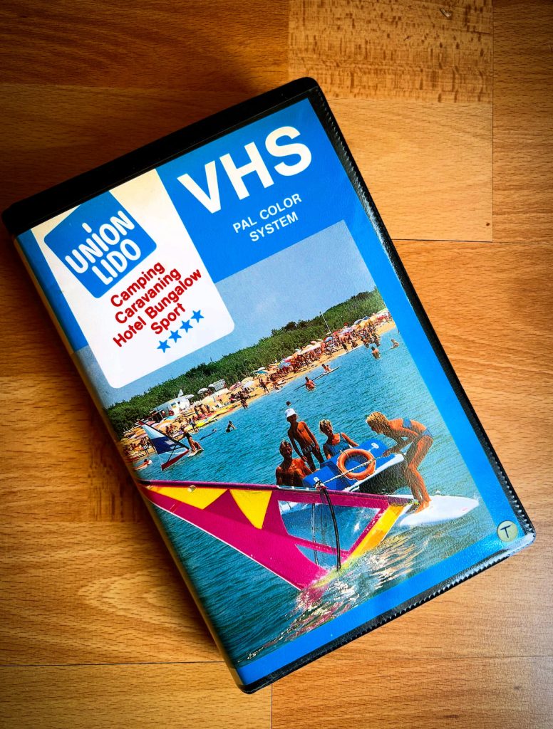 Bild 1 – Vorderseite der VHS-Kassette Deutsch: VHS-Videokassette von Union Lido mit einem Titelbild, das Wassersport auf dem Meer und den Strand mit Sonnenschirmen zeigt. Das Cover trägt das Union-Lido-Logo sowie die Aufschrift „Camping, Caravanning, Hotel, Bungalow, Sport“ und „VHS PAL Color System“.