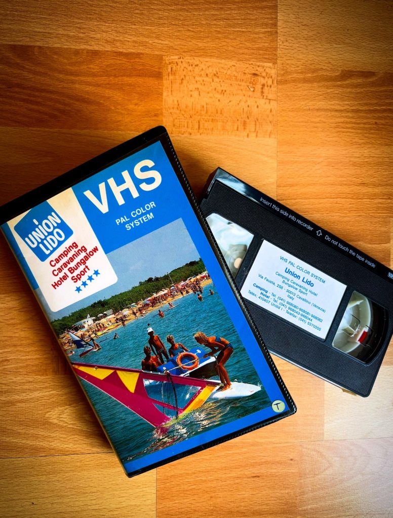 Union-Lido-VHS-Kassette neben ihrer Hülle auf einem Holztisch. Das Cover zeigt Wassersport auf dem Meer und den Strand; auf dem Videoband ist ein Etikett mit dem Namen Union Lido und den Kontaktdaten des Campingplatzes zu sehen.