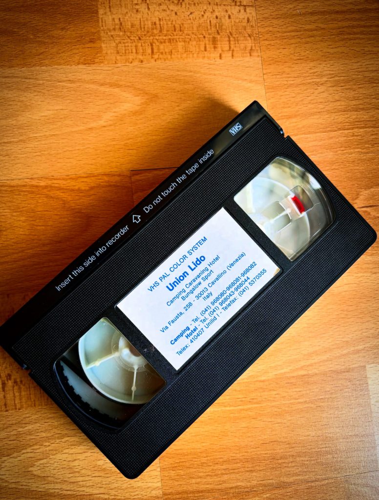 VHS-Videokassette von Union Lido mit schwarzem Gehäuse, sichtbaren Spulen und einem weißen Etikett mit dem Namen Union Lido sowie den Kontaktdaten des Campingplatzes in Cavallino, Italien.