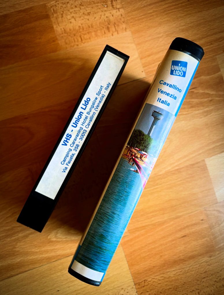 Seitliche Ansicht einer Union-Lido-VHS-Kassette und ihrer Hülle auf einem Holztisch. Auf dem Etikett der Hülle stehen der Name Union Lido, die Urlaubsangebote und die Adresse in Cavallino bei Venedig. Die Hülle zeigt außerdem einen Ausschnitt des Strandmotivs vom Cover.
