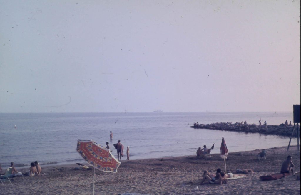 Historische Aufnahme des Strandes von Union Lido in Cavallino aus dem Jahr 1969 mit Badegästen, Sonnenschirmen, dem Meer und einem steinernen Wellenbrecher.