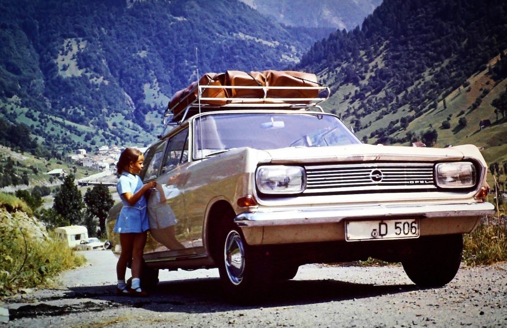 Historische Aufnahme der Anreise über den Brenner im Jahr 1966. Ein Opel Rekord mit Gepäck auf dem Dach steht vor einer alpinen Berglandschaft, während ein Mädchen neben dem Fahrzeug steht. Die Aufnahme zeigt die typische Urlaubsreise mit dem Auto über die Alpen in Richtung Italien.
