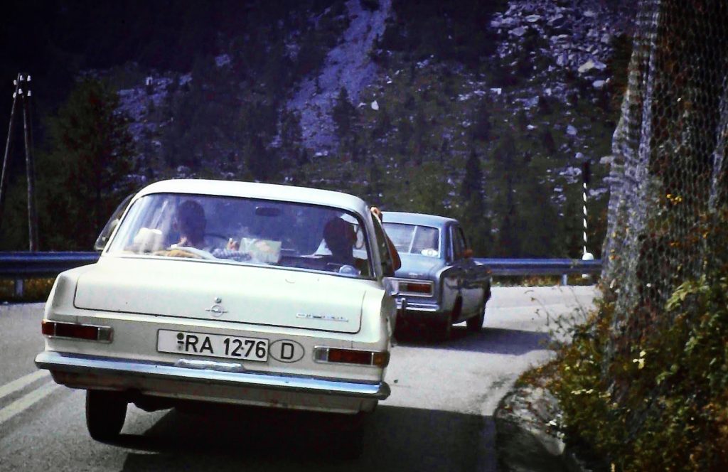 Historische Aufnahme der Anreise über den Brenner im Jahr 1966. Mehrere Opel-Fahrzeuge fahren auf einer Bergstraße durch die alpine Landschaft. Die Aufnahme dokumentiert die damalige Urlaubsreise mit dem Auto in Richtung Italien.